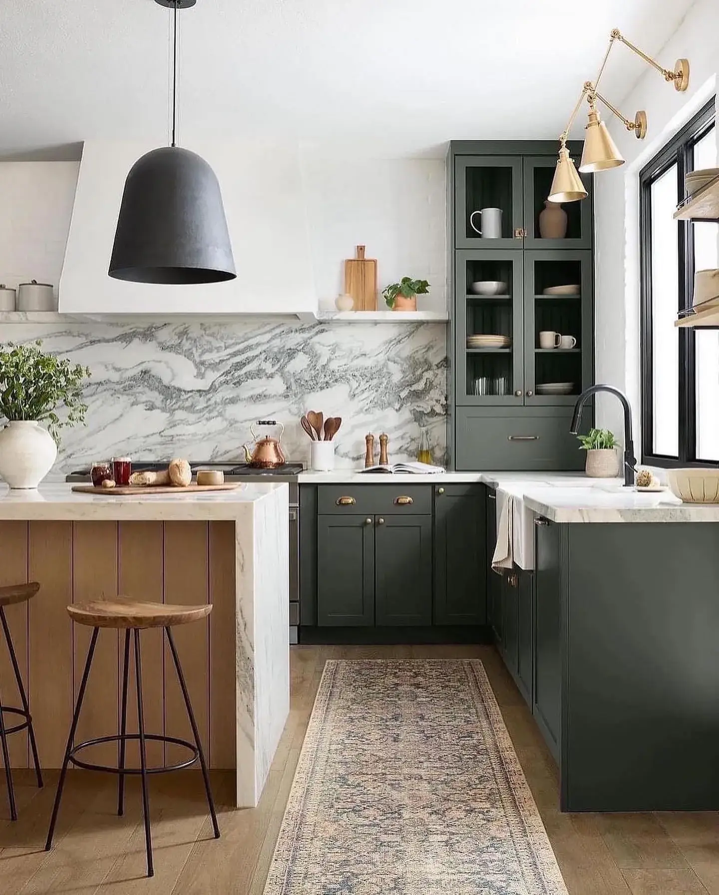 Kitchen remodel sink-side view with hunter-green cabinets, marble counters and backsplash, brass swing-arm sconces, and slatted oak island by Zona Floors
