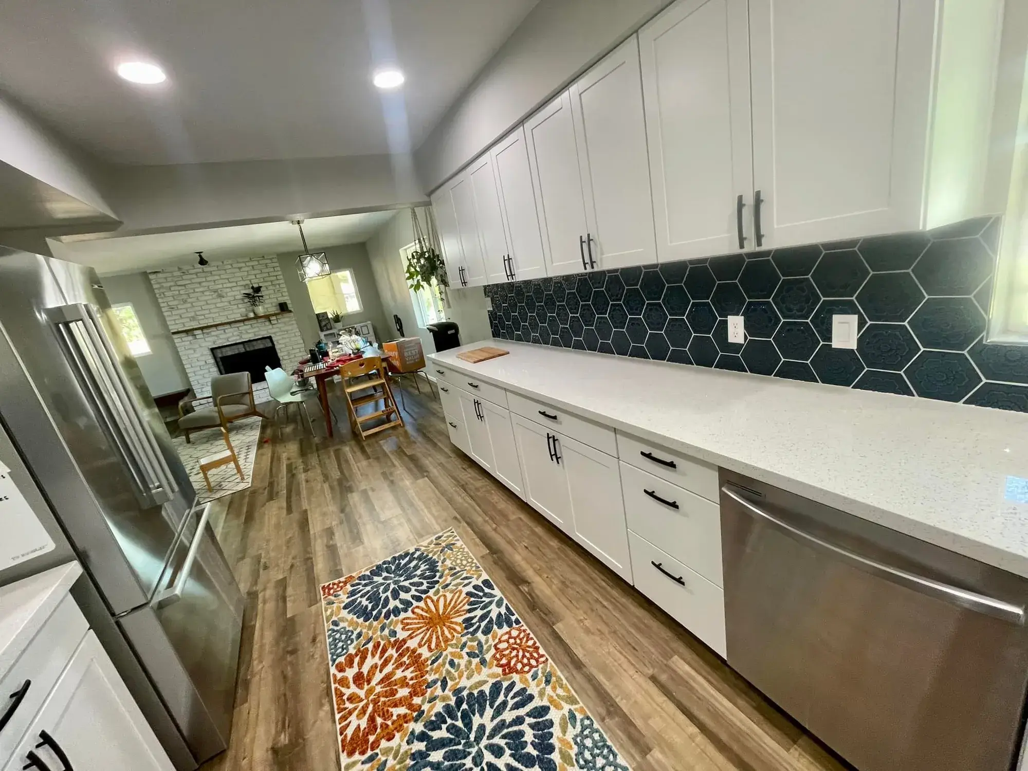 Completed kitchen remodel in Arizona by Zona Floors with white shaker cabinets, teal hexagon tile backsplash, quartz counters, and wood-look LVP flooring opening to a brick fireplace living room